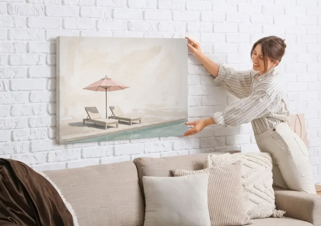 Two Chaise Longues Under a Pink Umbrella canvas wall art being hung on a white brick wall above a sofa
