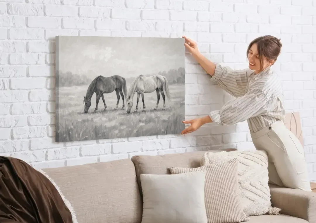 Two Horses Grazing in Misty Pasture in Black and White canvas wall art being hung on a white brick wall above a sofa