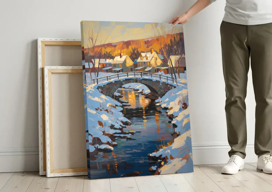 Winter Village Painting with Stone Footbridge canvas wall art leaning against a wall next to blank canvases